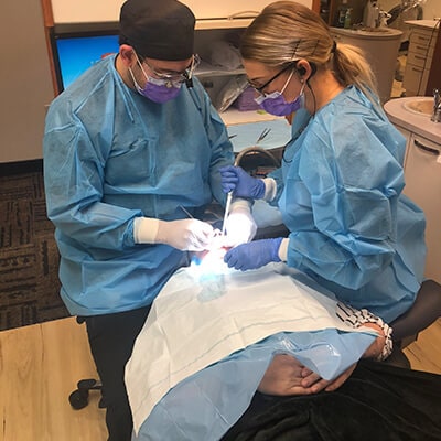 Dr. Neal Raval, in the surgery room operating a patient's mouth, while his dental assistant helps him, both wearing mouths and blue suits for surgery