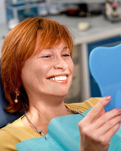 A mature woman looking at her new teeth in a blue mirror inside our Bellevue dental office.