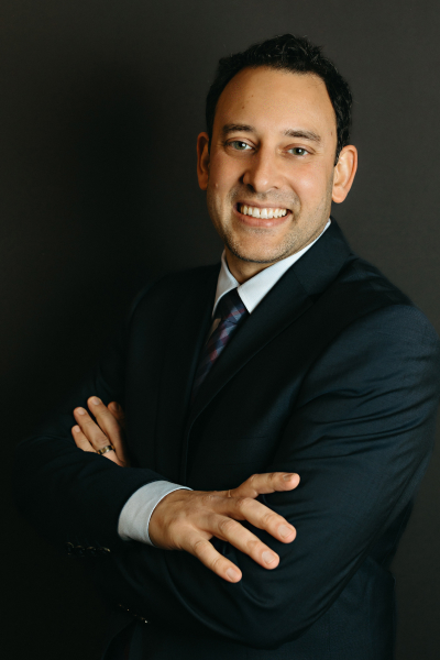 Dr. Neal Raval wearing a black suit with a white shirt, while smiling and wearing a black necklace 